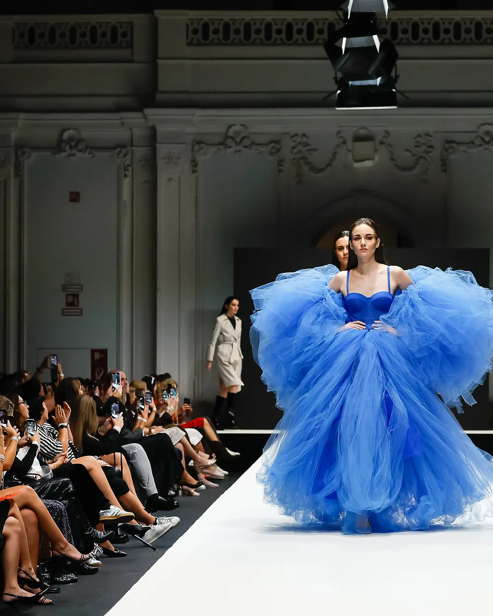 Model in blauer Tüllrobe, das Publikum sitzt dicht am Laufsteg