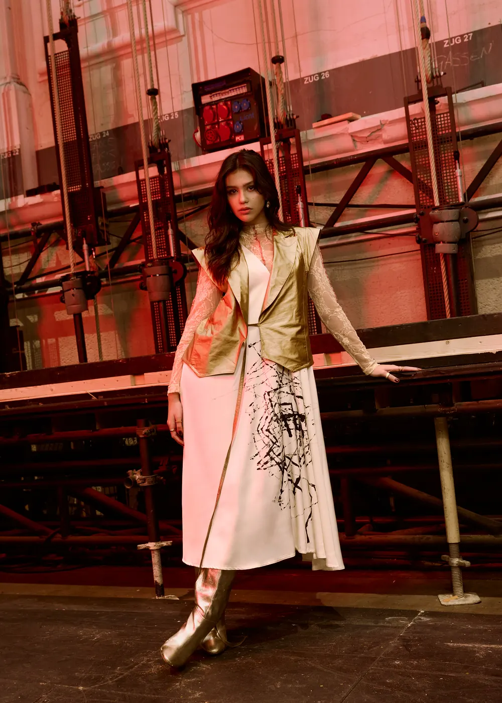 Model in goldener Weste über einem hellen Kleid vor der Bühnentechnik einer Halle